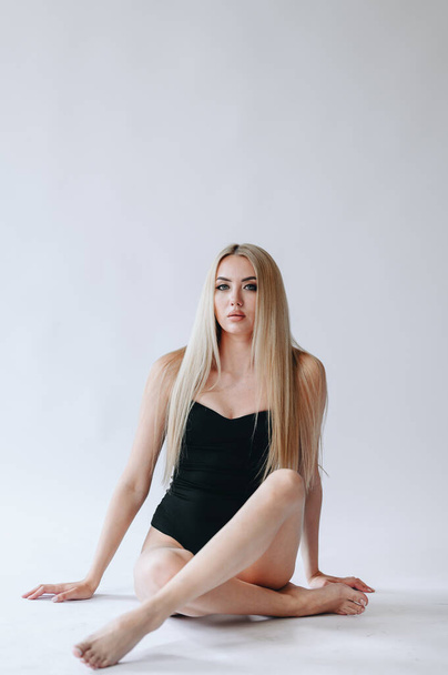Intimo pubblicitario.Foto verticale di una ragazza con i capelli lunghi si siede in un body nero su uno sfondo bianco a gambe incrociate - Foto, immagini