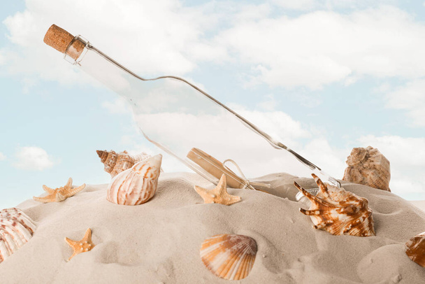 Corked glass bottle with rolled paper note and seashells on sand against sky - 写真・画像