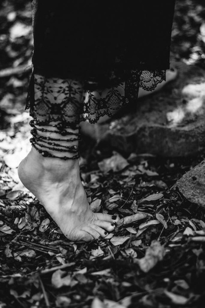 Beautiful feet of young gothic and witch woman with black nails and accessories walking over autumn leaves in the forest ground (in black and white) - Photo, Image