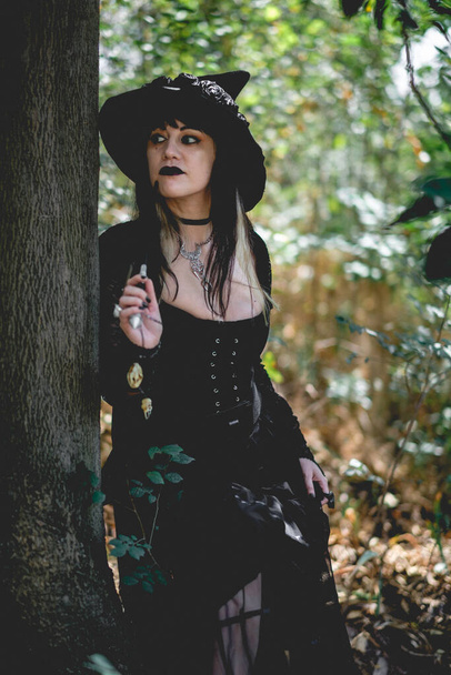 Hermosa mujer joven, gótica, sexy y bruja de manos estilizadas con uñas y accesorios negros, varita mágica de madera y sombrero de bruja en el bosque entre la naturaleza - Foto, imagen