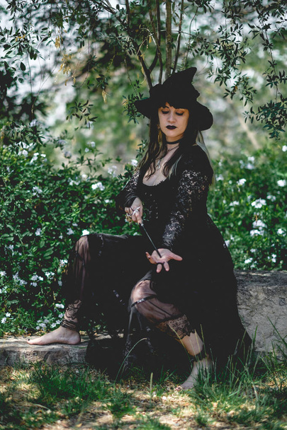 Beautiful young, gothic, sexy and witch woman from with stylized hands with black nails and accessories, magic wooden wand and witch hat in the forest between the nature - Photo, Image