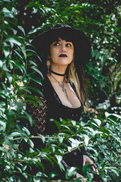 Schöne junge, gotische, sexy und Hexe Frau aus mit stilisierten Händen mit schwarzen Nägeln und Accessoires, Zauberstab und Hexenhut im Wald zwischen der Natur - Foto, Bild