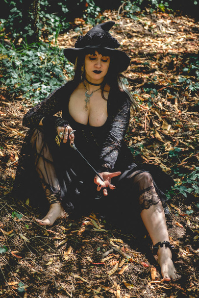 Güzel, genç, gotik, seksi ve cadı kadın. Tarzlanmış elleri, siyah tırnakları ve aksesuarları, sihirli ahşap değneği ve doğanın arasındaki ormanda cadı şapkası olan kadın. - Fotoğraf, Görsel