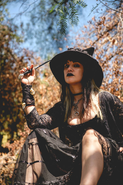 Beautiful young, gothic, sexy and witch woman from with stylized hands with black nails and accessories, magic wooden wand and witch hat in the forest between the nature - Фото, зображення