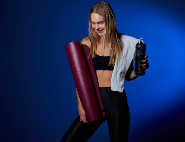 Chica deportiva, con una toalla en el hombro y una botella de agua en las manos, está descansando después de entrenar en un rincón oscuro del gimnasio - Foto, Imagen