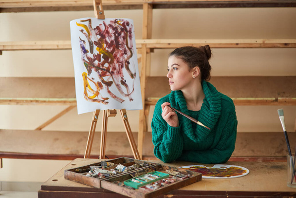 Esmer sanatçı kız elinde fırçalarla sehpanın yanında duruyor. Sanat atölyesinde yaratıcı bir ortam - Fotoğraf, Görsel