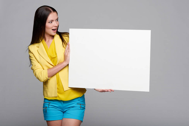 Surprised girl in yellow and blue, holding empty whiteboard, looking at blank copy space at board - Foto, Imagem