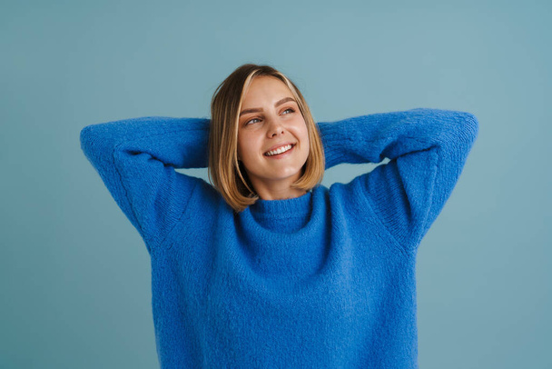 Young blonde woman smiling while posing with hands behind head isolated over blue background - Foto, Bild