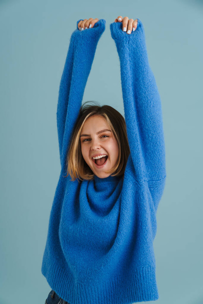 Νεαρή ξανθιά γυναίκα ουρλιάζει ενώ ποζάρει με τα χέρια ψηλά απομονωμένη πάνω από το μπλε φόντο - Φωτογραφία, εικόνα