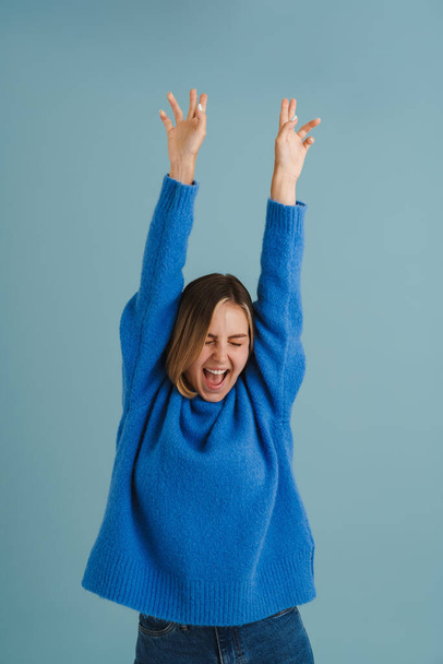 Young blonde woman screaming while posing with hands up isolated over blue background - Zdjęcie, obraz