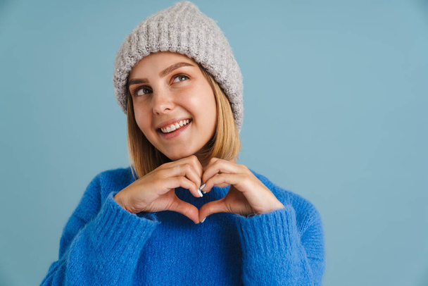 Young blonde woman wearing knit hat showing heart gesture isolated over blue background - Photo, Image