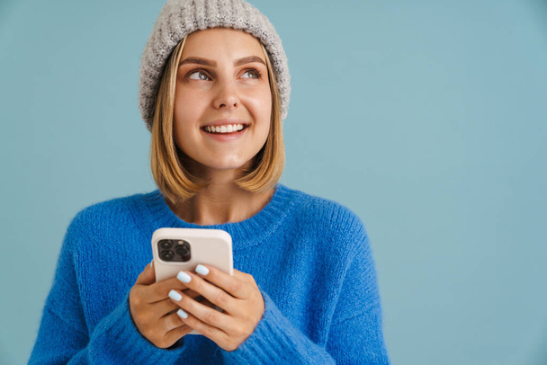 Young blonde woman wearing knit hat smiling and using cellphone isolated over blue background - Фото, изображение