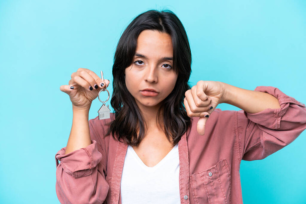 Junge hispanische Frau hält Hausschlüssel isoliert auf blauem Hintergrund und zeigt Daumen nach unten mit negativem Ausdruck - Foto, Bild