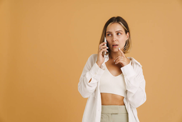 Young puzzlement woman talking on mobile phone isolated over beige background - Fotoğraf, Görsel