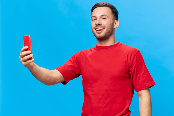Mosolygó cserzett jóképű férfi piros pólóban csinál videohívást telefonos nézés kamera pózol elszigetelt kék stúdió háttérben. Másold le a Banner Mockup-ot. Online emberek életmód koncepció - Fotó, kép