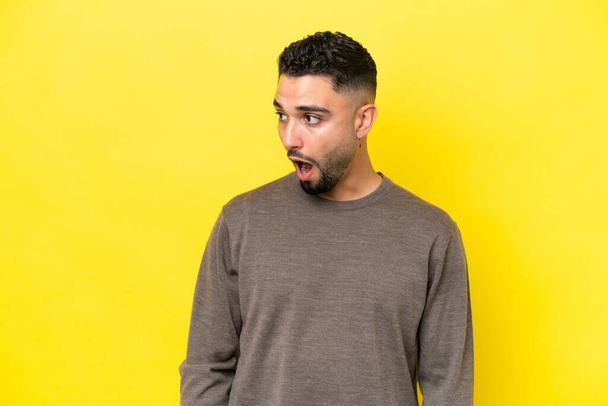 Young Arab handsome man isolated on yellow background doing surprise gesture while looking to the side - 写真・画像