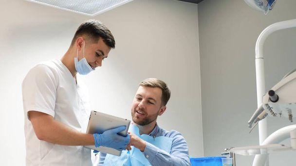 Der Zahnarzt verwendet eine Tablette, um dem Patienten Bilder der Zähne zu zeigen. Schmerzfreie Zahnbehandlung in einer modernen Zahnklinik. - Foto, Bild