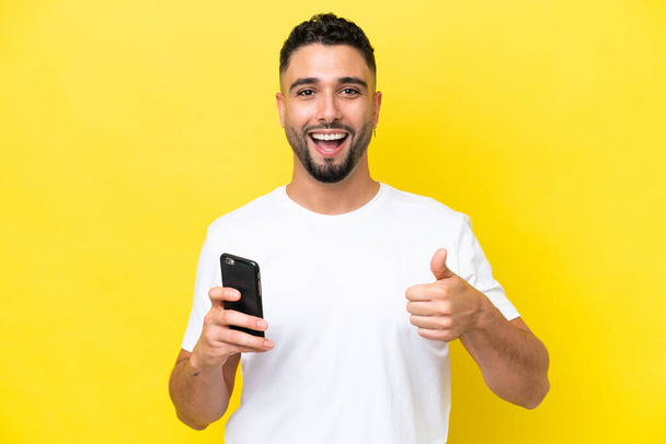 Young Arab handsome man isolated on yellow background using mobile phone while doing thumbs up - Foto, Imagen