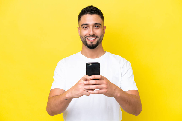 Young Arab handsome man isolated on yellow background looking at the camera and smiling while using the mobile - Fotó, kép