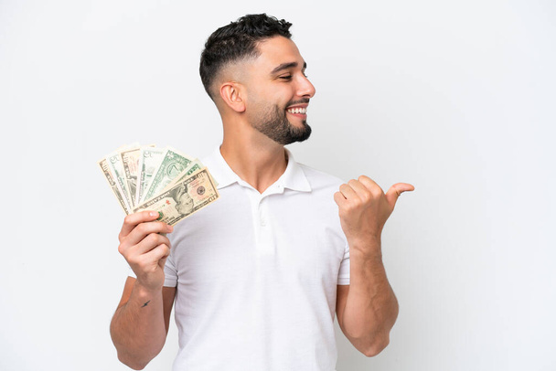 Young Arab man taking a lot of money isolated on white background pointing to the side to present a product - Foto, Imagen