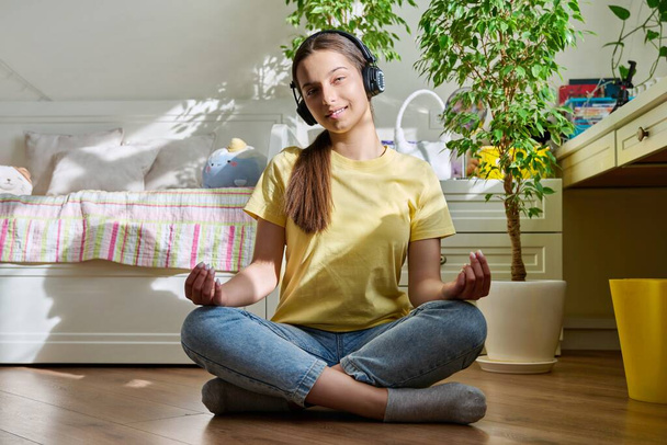 Teenage girl in headphones relaxing meditating sitting in lotus position at home on floor in room, smiling female looking at camera. Recreation, lifestyle, adolescence, lifestyle, youth concept - Fotoğraf, Görsel
