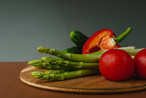 As verduras estão em uma tábua de madeira: tomates, espargos, pepinos, pimentos de sino vermelhos. marrom, fundo cinza escuro. lugar para texto - Foto, Imagem