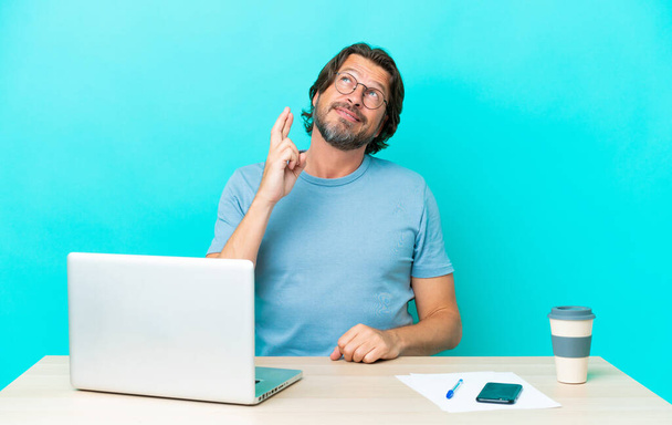 Vezető holland férfi egy asztalnál egy laptop elszigetelt kék háttérrel, ujjak keresztbe, és a legjobbakat kívánva - Fotó, kép