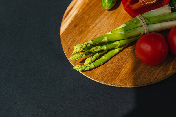 Las verduras se encuentran en una tabla de madera: tomates, espárragos, pepinos, pimientos rojos. marrón, fondo gris oscuro. lugar para el texto - Foto, Imagen