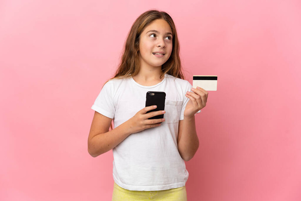 Child over isolated pink background buying with the mobile with a credit card while thinking - Fotó, kép