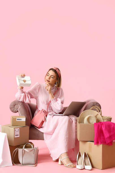 Nachdenkliche junge Frau mit Laptop und neuer Kleidung sitzt im Sessel auf rosa Hintergrund - Foto, Bild
