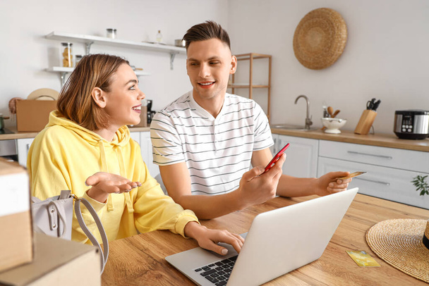 Mladý pár s notebookem, mobilním telefonem a nakupováním kreditních karet online v kuchyni - Fotografie, Obrázek