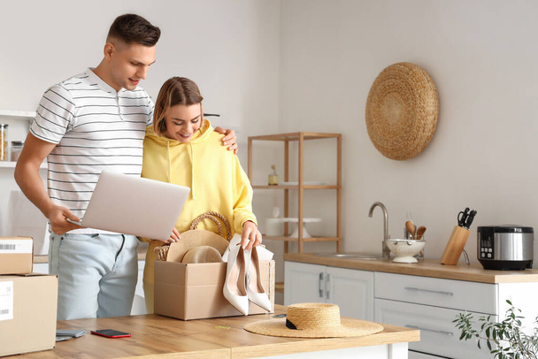 Young couple with laptop unpacking box with new clothes in kitchen. Online shopping - Фото, изображение