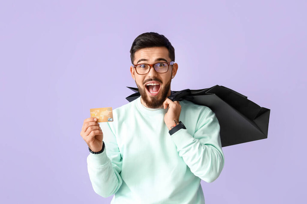 Szczęśliwy młody człowiek z karty kredytowej i torby na zakupy na tle liliowym - Zdjęcie, obraz