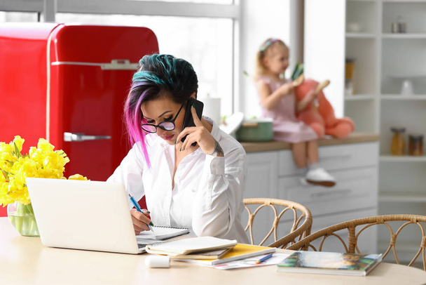 Matka pracuje z laptopem i telefonem komórkowym przy stole, podczas gdy jej córeczka bawi się w kuchni - Zdjęcie, obraz