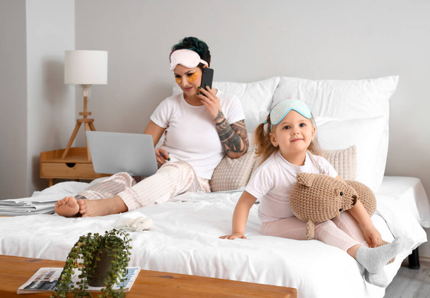 schattig klein meisje met speelgoed en haar werkende moeder in slaapkamer - Foto, afbeelding