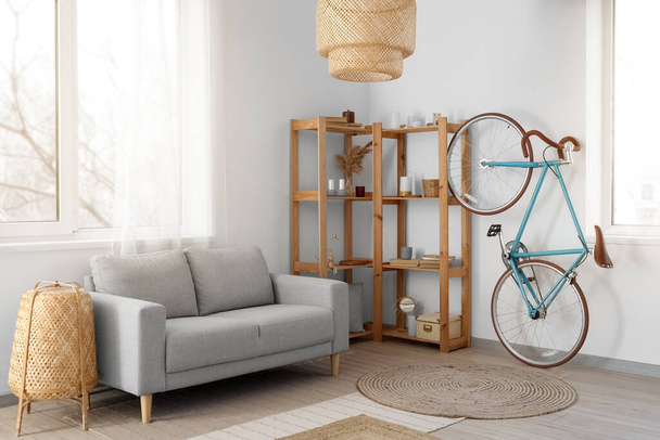 Интерьер светлой гостиной с велосипедом, серым диваном и деревянными полками - Фото, изображение