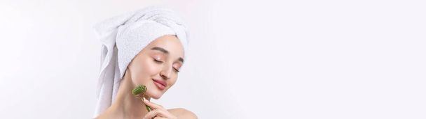 Beautiful girl with thick eyebrows and perfect skin at white background, towel on head, beauty photo. Holds a cosmetic roller. - Фото, изображение