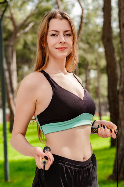 Portrait of young brunette woman wearing sports bra standing on city park, outdoors flat stomach with a skipping rope in her hands at waist level. Healthy life, outdoor sport concepts. - Foto, afbeelding