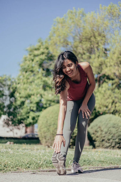 Привлекательная улыбающаяся индийская молодая бегунья растягивает мышцы телят перед тем, как побежать в парк - Фото, изображение