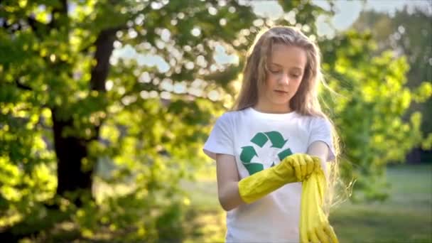 Menina vestindo luvas de borracha para coleta de lixo de plástico em um parque poluído, sinal de reciclagem na camiseta. Movimento lento - Filmagem, Vídeo