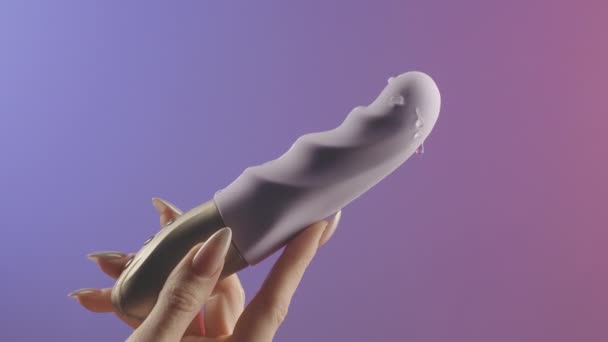 Κοντινό πλάνο της γυναίκας χέρι κρατώντας θηλυκό παιχνίδι σεξ. Δημιουργικό. Υγρό διαφανές λιπαντικό που πέφτει σε ένα σύγχρονο δονητή απομονωμένο σε ροζ και μοβ φόντο κλίση - Πλάνα, βίντεο