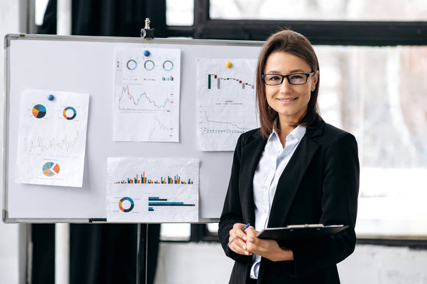 Portrait of successful young adult pretty, confident caucasian woman, financial top manager, wearing business suit and glasses, standing in a modern office near markerboard, looks at camera, smiling - Photo, Image