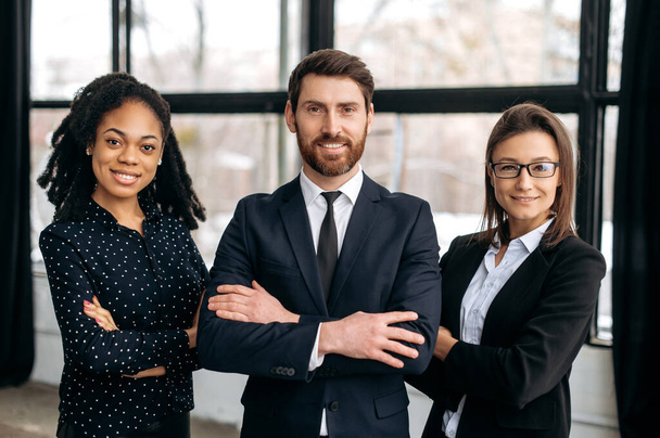 Group portrait of business partners. Multiracial successful colleagues, dressed in formal wear, standing in their modern office with their arms crossed, looking at the camera, smiling friendly - Photo, Image