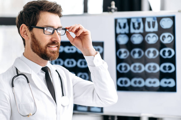 Foto di un medico generico maschio caucasico professionista intelligente, chirurgo, polmonologo, in uniforme medica, con occhiali e stetoscopio sulle spalle, in piedi in clinica, distoglie lo sguardo, sorride - Foto, immagini