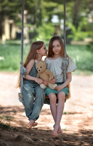 Happy laughing kids girls sisters with long hair enjoying a swing ride with a teddy bear toy on a playground in a park on a sunny summer day - Foto, imagen