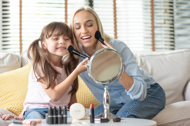 Attractive Happy Smiling Mother and little Daughter looking at the mirror and applying cosmetic with brushes on the cheekbones. - Foto, afbeelding