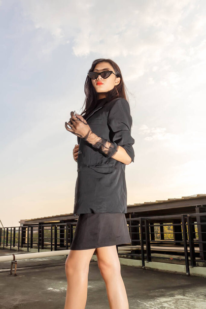 Concepto de moda, mujer de moda posando en vestido negro con gafas de sol mientras está de pie en la azotea. - Foto, Imagen