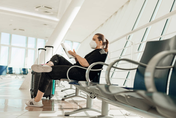 Вид сбоку женщины в черной одежде, белой одноразовой маске, сидящей на стуле возле чемоданов в транзитном зале аэропорта, держащей журнал, ожидающей полета самолета. Безопасность личного здоровья. - Фото, изображение