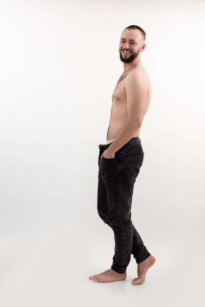Vertical confident and cool attractive man with beard, smiling. Shirtless and topless posing with expression in studio on white background. Body positive beauty side view. Full body, hands in pockets - Photo, image