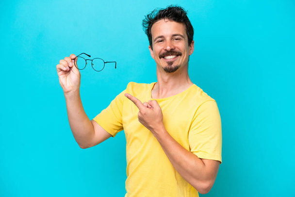 Jeune homme caucasien tenant des lunettes isolées sur fond bleu et le pointant - Photo, image
